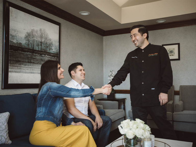 Three people are in a warmly decorated room. A person in a black jacket shakes hands with someone seated on a couch in a denim jacket and yellow skirt. Another person in a white top and jeans sits nearby, creating a welcoming, conversational atmosphere amid modern furnishings and floral decor