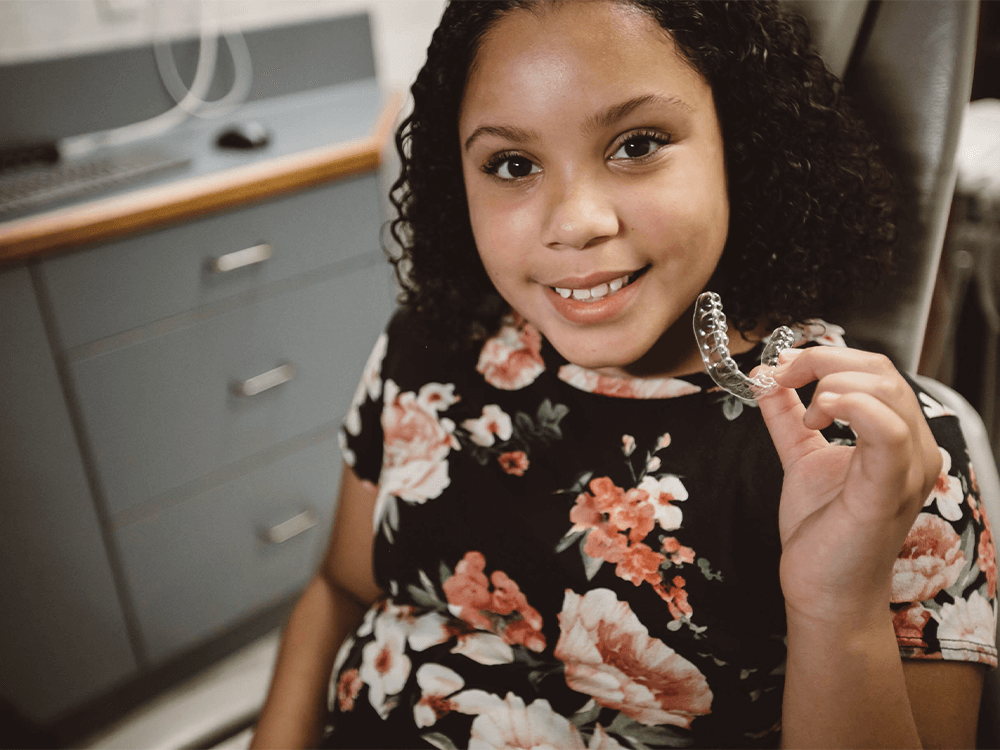 A person in a dental office holds a clear dental aligner in their hand. They wear a black floral-patterned shirt. Background includes a counter, drawers, and dental equipment.
