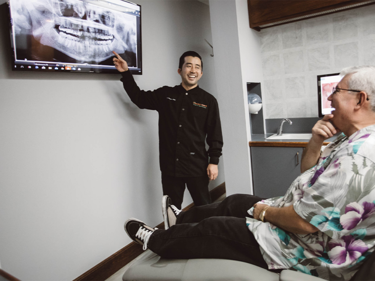 Dr. Nishimura in a black uniform points at a dental X-ray displayed on a monitor while explaining the results to a seated patient. The office features a dental chair, sink, and cabinets, creating a typical consultation setting.