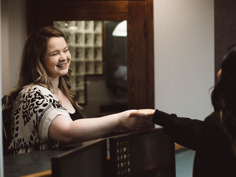 Two people shaking hands across a counter, possibly in a professional or service-related setting, with a wooden and glass background.